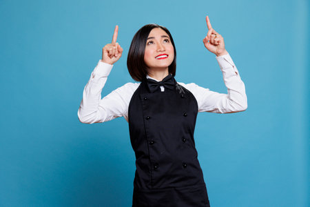 Young asian waitress wearing uniform looking and pointing upwards with fingers, promoting product. Restaurant cheerful woman employee showing up in studio on blue backgroundの写真素材