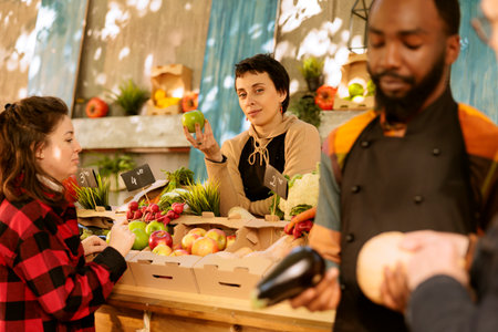 Young woman with an apron working at farmers market booth, offering fresh organic fruits and veggies. Female farmer grasping an apple and looking at camera while standing behind harvest fair stall.の写真素材