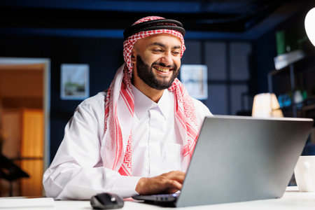 Enthusiastic Middle Eastern businessman efficiently works on his laptop while seated at his home office desk. Profile shot of Arab guy checking e-mail, surfing the net, and taking detailed notes.の写真素材