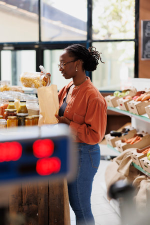 Fusilli pasta is poured into a brown paper bag by black woman wearing glasses in grocery store. African American lady fills plastic free bag with freshly made spaghetti at bio food convenience shop.の写真素材