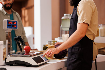 Caucasian storekeeper wearing black apron, weighs a red apple on digital scale and checks the value, while male customer waits. Salesman assisting client with measuring his bio food products.の写真素材