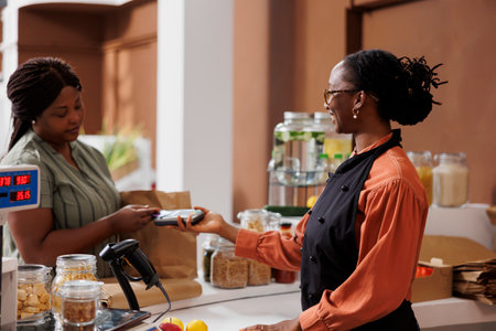 African American woman happily makes cashless payment at market counter using credit card. She smiles as she communicates with the vendor and completes the transaction with a debit and pos machine.の写真素材