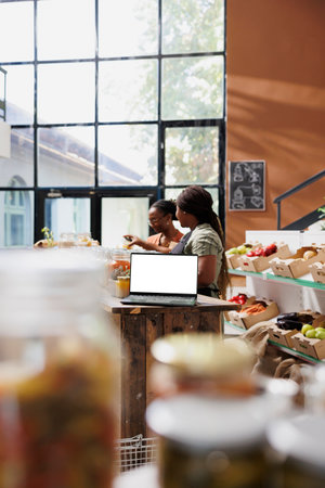 Laptop on counter shows blank copyspace mockup template as the seller helps client in eco friendly shop. In bio food store selling healthy goods, isolated screen device functions as promotional tool.の写真素材