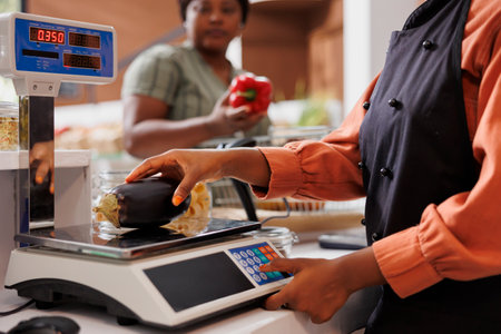 African american cashier weighing locally grown eggplant for female customer at the counter. Shopkeeper utilizing digital scale to measure weight of freshly harvested bio vegetable.の写真素材