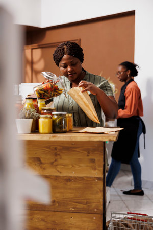 African American woman shopping for fresh organic produce and local products in a sustainable supermarket. Portrait of female customer pouring fresh pasta into brown paper bag.の写真素材