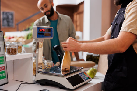 At checkout counter caucasian vendor places brown paper bag on weighing scale to check value for male customer. Arab man waiting at cashier desk while shopkeeper measures his eco friendly parcel.の写真素材