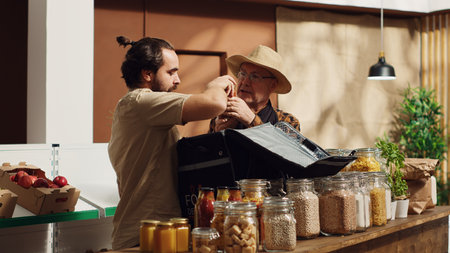 Deliveryman providing organic zero waste supermarket food orders to customers, helped by elderly storekeeper to fill thermic backpack. Man bringing local neighborhood shop groceries to clientsの写真素材