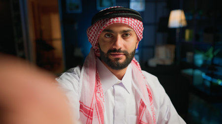 POV of cheerful Muslim content creator dressed in traditional clothing filming himself. Smiling Arab man with thobe and headscarf waving towards camera, doing online vloggingの写真素材