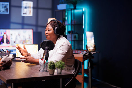Female celebrity is seen answering interviewer questions in home studio, utilizing smartphone on tripod to record encounter. Black woman filming content for online audience, sharing exciting stories.の写真素材
