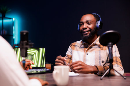 Black guy with a notebook is using podcast equipment to provide online advice. Male African American vlogger is seated at the table, recording an episode at home while speaking into a microphone.の写真素材