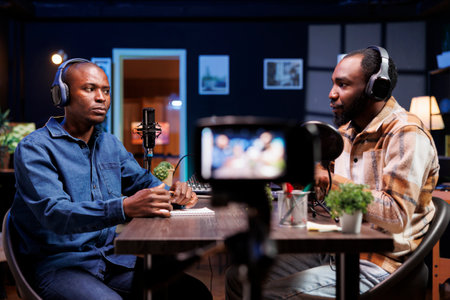 Content creators sitting at table, recording video material with professional camera at home. Online show host conducts friendly chat with media star while filming with device mounted on tripod.の写真素材