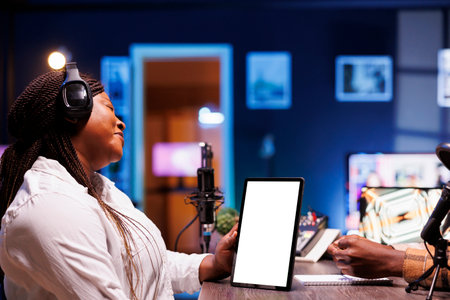 Black woman grasping device displaying blank white screen while hosting a talk show. Female content creator using headphones and podcast equipment while holding tablet with chromakey template.の写真素材