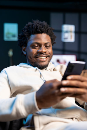 Cheerful man impatiently texting friend, discussing meeting hour. Person sitting on cozy sofa in modern stylish apartment typing on mobile phone, excited to meet up with mateの写真素材
