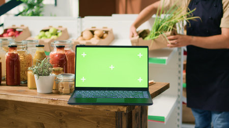 Laptop displaying greenscreen in bio shop filled with freshly harvested fruits and vegetables, sustainable lifestyle concept. Computer shows isolated chromakey template in zero waste store.の写真素材