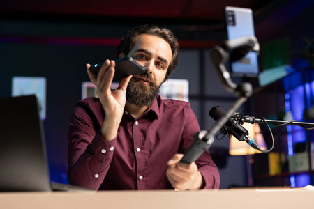Tech expert assessing smartphone performance, talking about components used inside, filming with selfie stick. Entertainer unpacking mobile phone to review it on technology internet channelの写真素材
