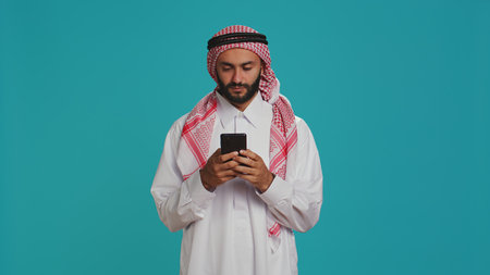 Islamic man works on his mobile gadget, surfing through social media platforms or online pages. Middle eastern guy in customary apparel uses smartphone to access global web, feeling excited.の写真素材