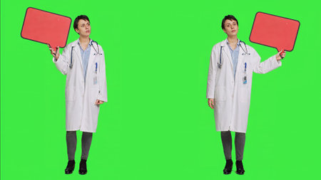 Woman doctor holding a red empty speech bubble sign in studio, showing an isolated blank cardboard icon against greenscreen backdrop. Physician creating announcement with banner. Camera A.の写真素材