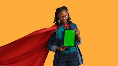 Portrait of joyful woman portraying superhero showcasing green screen tablet, giving positive feedback, studio background. Radiant person doing hero cosplay recommending mockup device, camera Bの写真素材