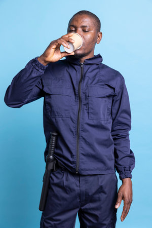 African american agent drinking coffee cup against blue background, serving caffeine refreshment to get more energy after a sleepless night. Male security guard poses in front of camera.の写真素材