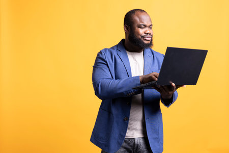 Smiling african american man looking at business diagram and figures on laptop, isolated over studio background. Upbeat worker checking company financial annual report charts on notebookの写真素材