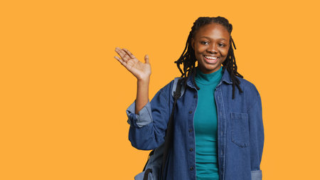 Portrait of chipper girl holding backpack, waving hand, gesturing, isolated over yellow studio background. Jolly african american student with school rucksack doing greeting hand gesture, camera Aの写真素材