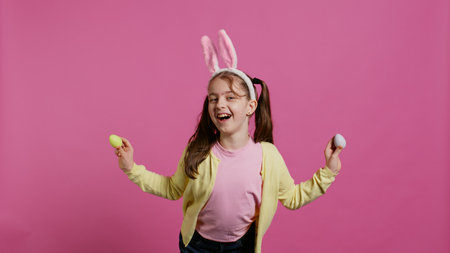 Adorable little girl playing peek a boo game in studio, showing her handcrafted colored easter eggs against pink background. Cheerful playful kid with bunny ears fooling around. Camera B.の写真素材