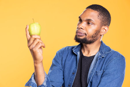 African american zero waste enthusiast looks at green apple on camera, enjoying raw healthy nutrition with ethically sourced fresh fruits. Advertising additives free eco friendly produce.の写真素材