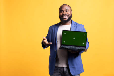 Portrait of radiant man holding green screen laptop, doing endorsement. Cheerful BIPOC person presenting chroma key notebook, doing positive assessment, isolated over studio backgroundの写真素材