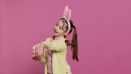 Lovely little girl with bunny ears sending air kisses in studio, presenting a stuffed pink rabbit toy for easter holiday. Smiling energetic preteen feeling positive and carefree. Camera A.の写真素材