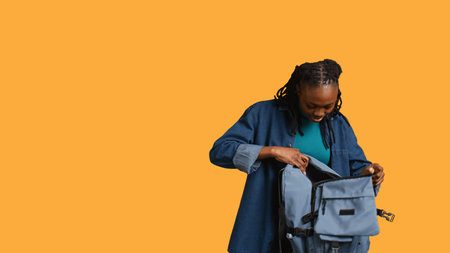 Joyous african american woman rummaging through backpack, looking for school notebooks, isolated over studio background. Merry teenager taking out university supplies out of rucksack, camera Bの写真素材