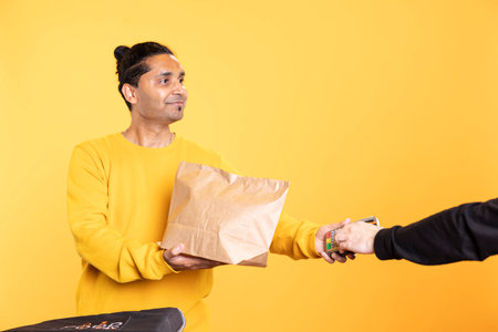 Jolly delivery worker offering groceries order to man paying with debit card. Customer buying meal from upbeat courier, swiping card on POS device, isolated over studio backdropの写真素材