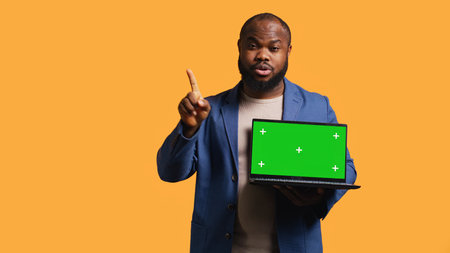 Upset african american man shows thumbs down sign gesturing holding green screen laptop, disapproving. Reviewer does rejection hand gesture regarding chroma key notebook, studio background, camera Bの写真素材