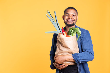 Happy young adult carrying a paper bag with raw organic produce, shopping for groceries at local farmers market. Man feels excited about ethically sourced food, zero waste bio concept.の写真素材