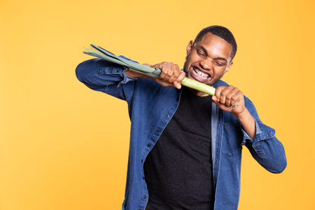 Portrait of model pretending to bite on a leek produce in studio, posing against yellow background with a green onion from grocery store. African american carefree guy promotes bio nutrition.の写真素材