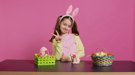 Cheery innocent child showing peace sign in studio and wearing bunny ears, celebrating easter sunday holiday with festive ornaments on a table. Happy energetic girl with creativity. Camera B.の写真素材