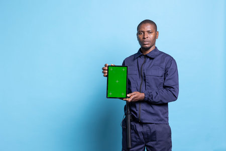 Mall security guard showing a tablet with isolated copyspace display on camera, wearing duty uniform equipped with baton and posing against blue background. Bodyguard with greenscreen.の写真素材