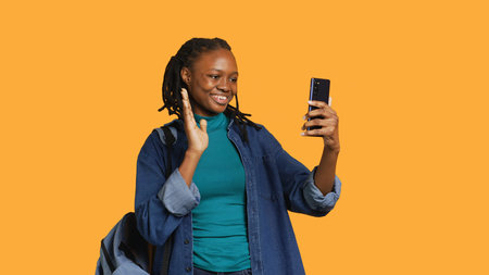 Cheerful woman saluting friends during teleconference meeting using smartphone, studio background....