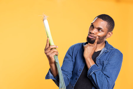 Pensive person inspecting a freshly harvested leek plant in studio, looking closely at green onion and promoting sustainable lifestyle. African american vegan guy examines fresh produce.の写真素材