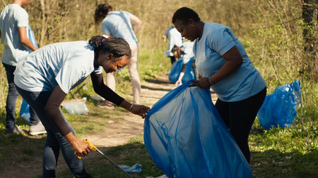 African american team of volunteers picking up trash from the ground, collecting rubbish and recycling in bags. Women cleaning the forest environment from plastic waste, illegal dumping. Camera A.の写真素材