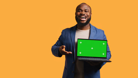 Smiling man holding isolated screen laptop, doing recommendation, talking with viewers. Cheerful BIPOC person pointing towards chroma key notebook, studio background, camera Bの写真素材