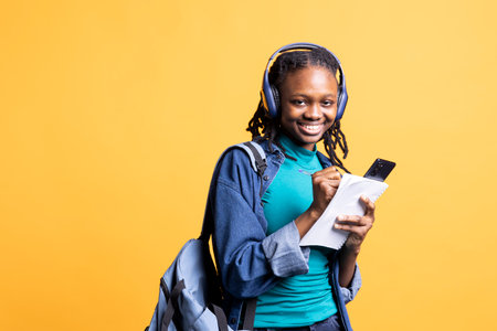 Portrait of smiling student writing homework, doing research online on smartphone and listening music, studio background. Jolly girl taking notes for school assessment, enjoying songs on headphonesの写真素材