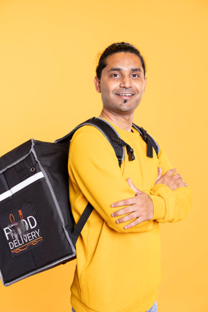Portrait of man holding thermal backpack, crossing arms, ready to fulfill orders, studio backdrop. Joyous food delivery service worker prepared to do delivery to customersの写真素材