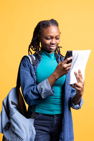 BIPOC young girl taking photos of school homework to send to colleague, isolated over studio background. Student photographing papers with information needed for university courses using phoneの写真素材