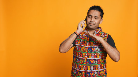 Happy man in traditional clothing chatting with best friend over telephone call. Upbeat person with colorful attire communicating with mate using phone, studio background, camera Bの写真素材