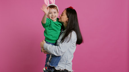 Mother lifting up her little boy and kissing him in studio, showing love and hugging cute kid against pink background. Sweet mom and son embracing each other and laughing. Camera B.の写真素材