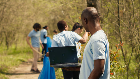 African american man looks at mockup screen on laptop while he participates in collecting rubbish from the forest area. Environmental activist works to protect the ecosystem. Camera A.の写真素材