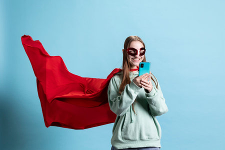 Smiling girl with mask portraying superhero, texting friends on mobile phone, studio background. Happy woman posing as hero in costume with mantle sending messages on smartphoneの写真素材