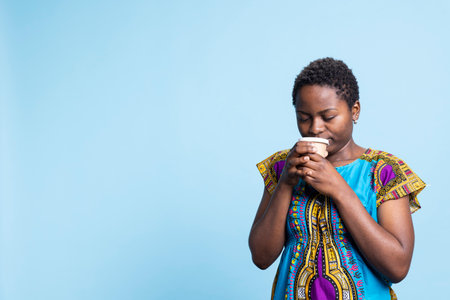 African american girl serving cup of coffee in front of camera, enjoys drink with natural caffeine aroma. Positive young adult drinking hot refreshment, wearing cultural bright attire over backdrop.の写真素材