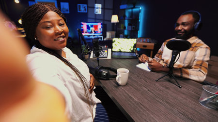 Woman using smartphone selfie camera to film podcast with guest telling interesting story to fans. African american man invited in studio sharing facts, filmed by mobile phone, POV shotの写真素材