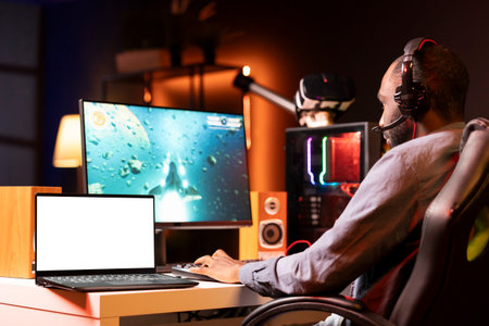 Mockup laptop on desk next to man playing SF videogame on computer, talking into headset with...